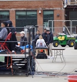 A group of people are gathered around an individual wearing a sports jersey with the number 11. They appear to be in a sports facility area, possibly a training or medical setup. There are tables, chairs, and equipment scattered around, with a brick building in the background. Some individuals are seated on a platform with electronics close by, suggesting a recording or monitoring activity. Utility vehicles and an orange barrier are present in the background.