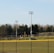 A sports field with a group of people engaging in athletic activities in the background. There is sports equipment like a soccer goal and a scoreboard visible. A chain-link fence runs across the foreground, and a statue or sculpture resembling an eagle is also seen. Trees and tall floodlights are positioned behind the field.