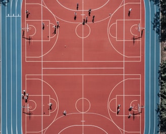 An aerial view of a large, vibrant sports complex featuring a red basketball court with multiple hoops and a surrounding blue running track. Several people are scattered across the area, engaged in various activities.