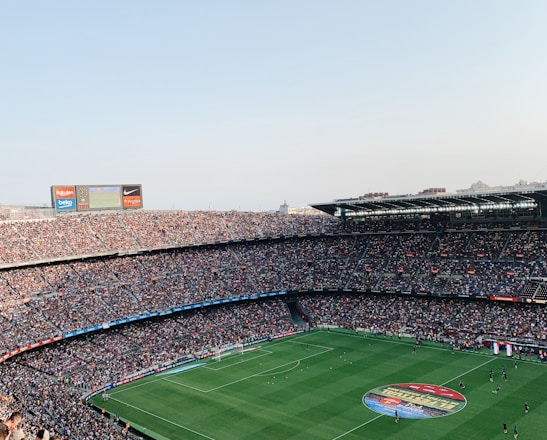 An expansive soccer stadium filled with thousands of spectators. The green field in the center features players and markings, surrounded by vibrant stands and advertising boards on the upper sides of the stadium. The audience is actively watching the game.