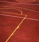 A sports court with a reddish-brown surface and bright yellow and white lines. The lines form geometric shapes, including rectangles and circles, suggesting markings for games like basketball or netball.