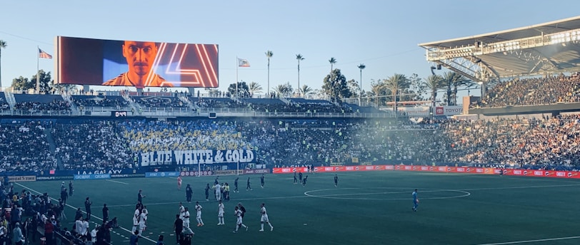 A large sports stadium filled with spectators. The field is occupied by soccer players, some walking and others scattered across the pitch. A massive screen displays a person, and flags fly on top of the stadium roof. The audience is dense, and the atmosphere seems lively with a visible banner showing 'BLUE WHITE & GOLD'.