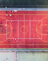 An aerial view of a sports court with a vibrant red surface and yellow lines marking different sections for basketball and soccer. Several people are present on the court, some engaged in activities, while others are standing along the perimeter. The court is enclosed by a metal fence and surrounded by grass on all sides.