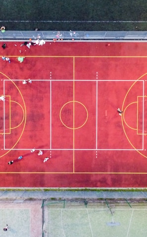 An aerial view of a sports court with a vibrant red surface and yellow lines marking different sections for basketball and soccer. Several people are present on the court, some engaged in activities, while others are standing along the perimeter. The court is enclosed by a metal fence and surrounded by grass on all sides.