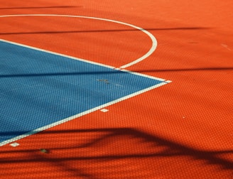 A vibrant sports court with sections of red and blue, featuring crisp white lines marking the boundaries. Shadows from a structure create geometric patterns across the surface, adding depth and interest to the scene.
