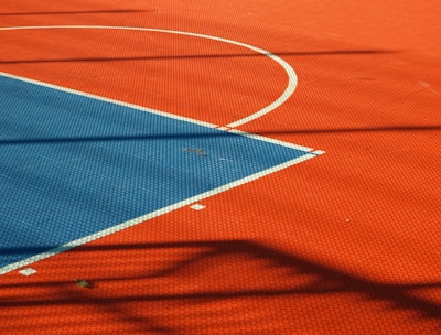 A vibrant sports court with sections of red and blue, featuring crisp white lines marking the boundaries. Shadows from a structure create geometric patterns across the surface, adding depth and interest to the scene.