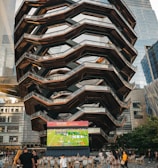 A large, spiral architectural structure with multiple levels stands prominently in an urban setting. In front of the structure, a large screen displays a sports broadcast, and several people are gathered around, some sitting and others standing. The scene is set outdoors, with towering modern buildings surrounding the area. The ground is paved with a geometric pattern of gray and white tiles.