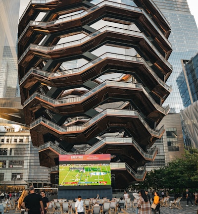 A large, spiral architectural structure with multiple levels stands prominently in an urban setting. In front of the structure, a large screen displays a sports broadcast, and several people are gathered around, some sitting and others standing. The scene is set outdoors, with towering modern buildings surrounding the area. The ground is paved with a geometric pattern of gray and white tiles.